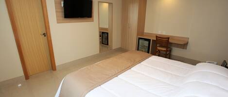 A bedroom with a large bed, a television mounted on the wall, a wooden wardrobe, and a desk with a chair.