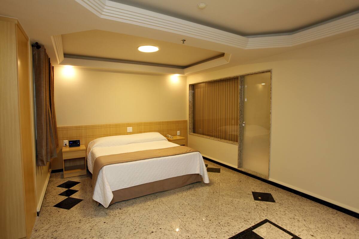 A hotel room with a bed, bedside tables, a window with blinds, and a tiled floor.