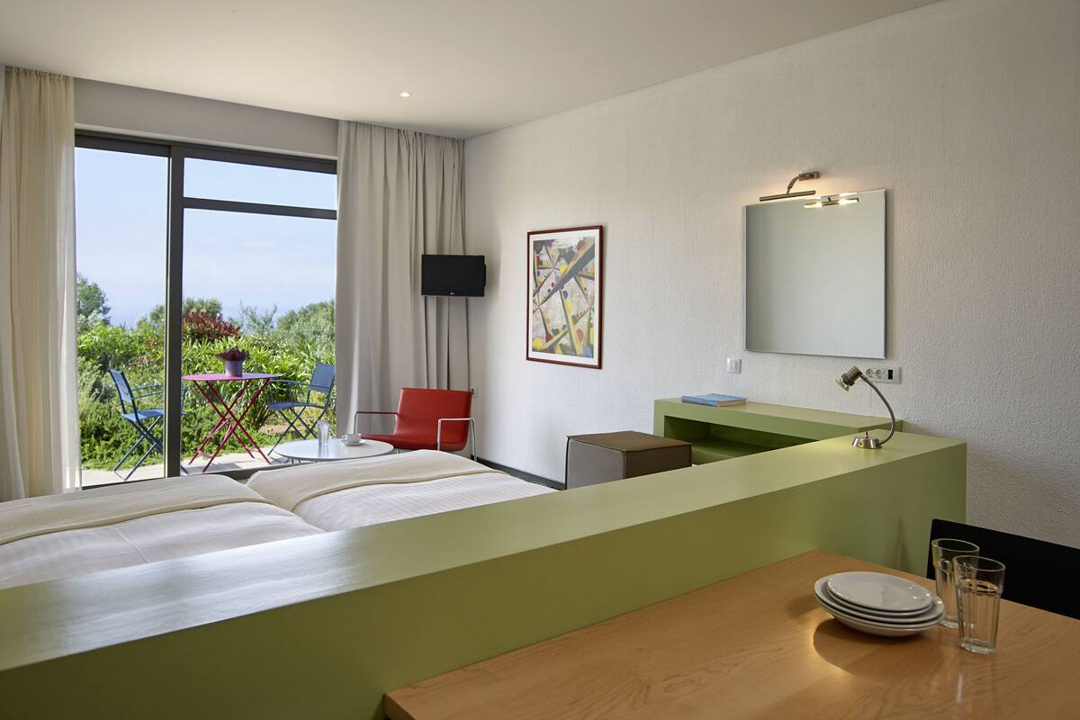 Habitación de hotel con cama, escritorio, silla, televisor, un cuadro y baño con espejo y lavamanos.