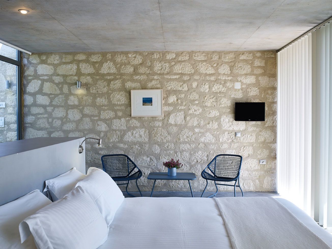 A hotel room with a stone wall, a bed, two chairs, a small table with a vase, and a TV mounted on the wall.