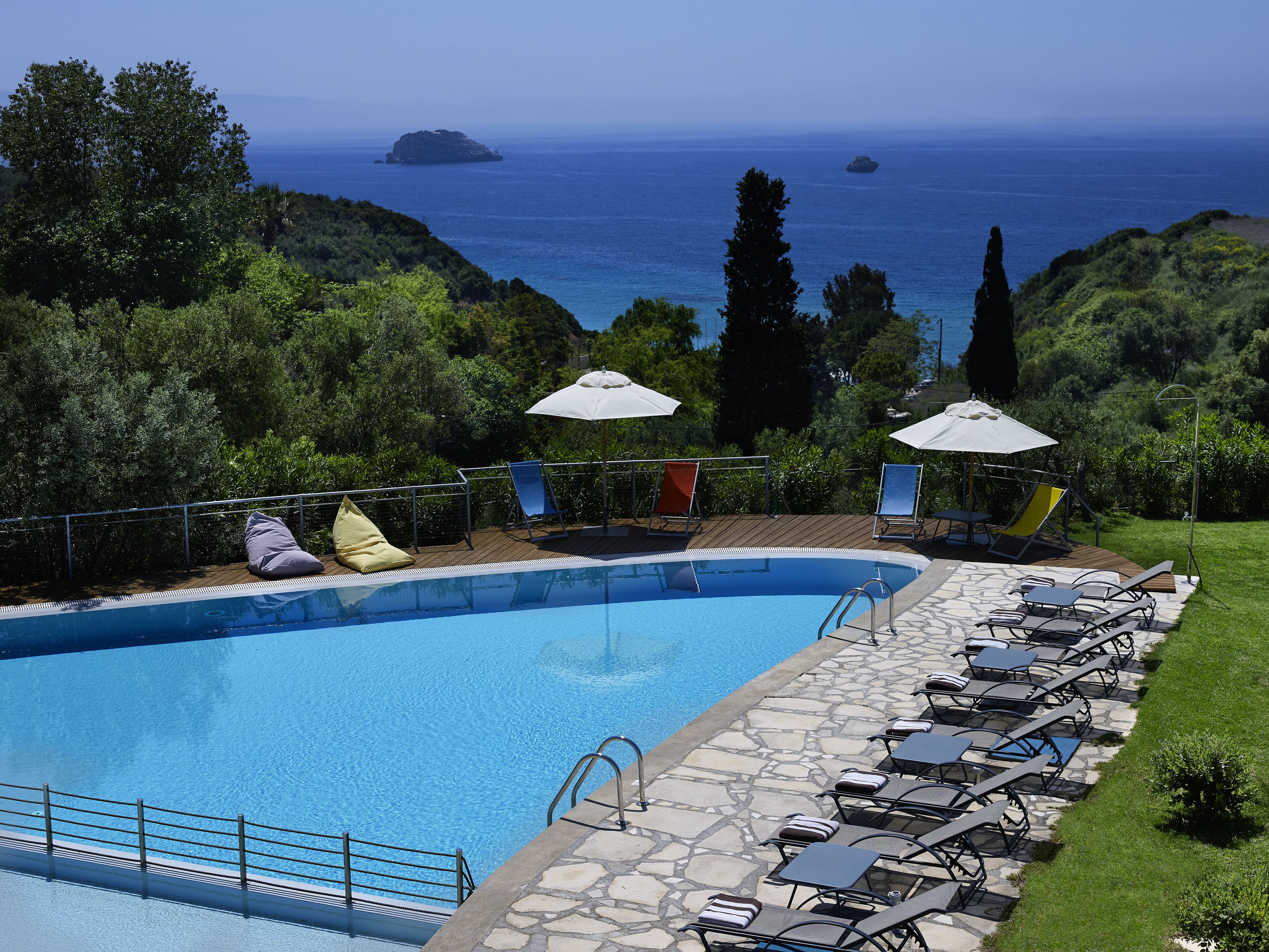 Ein Pool mit Liegestühlen und Sonnenschirmen, umgeben von viel Grün mit Blick auf das Meer.