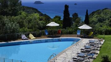 Ein Pool mit Liegestühlen und Sonnenschirmen, umgeben von viel Grün mit Blick auf das Meer.