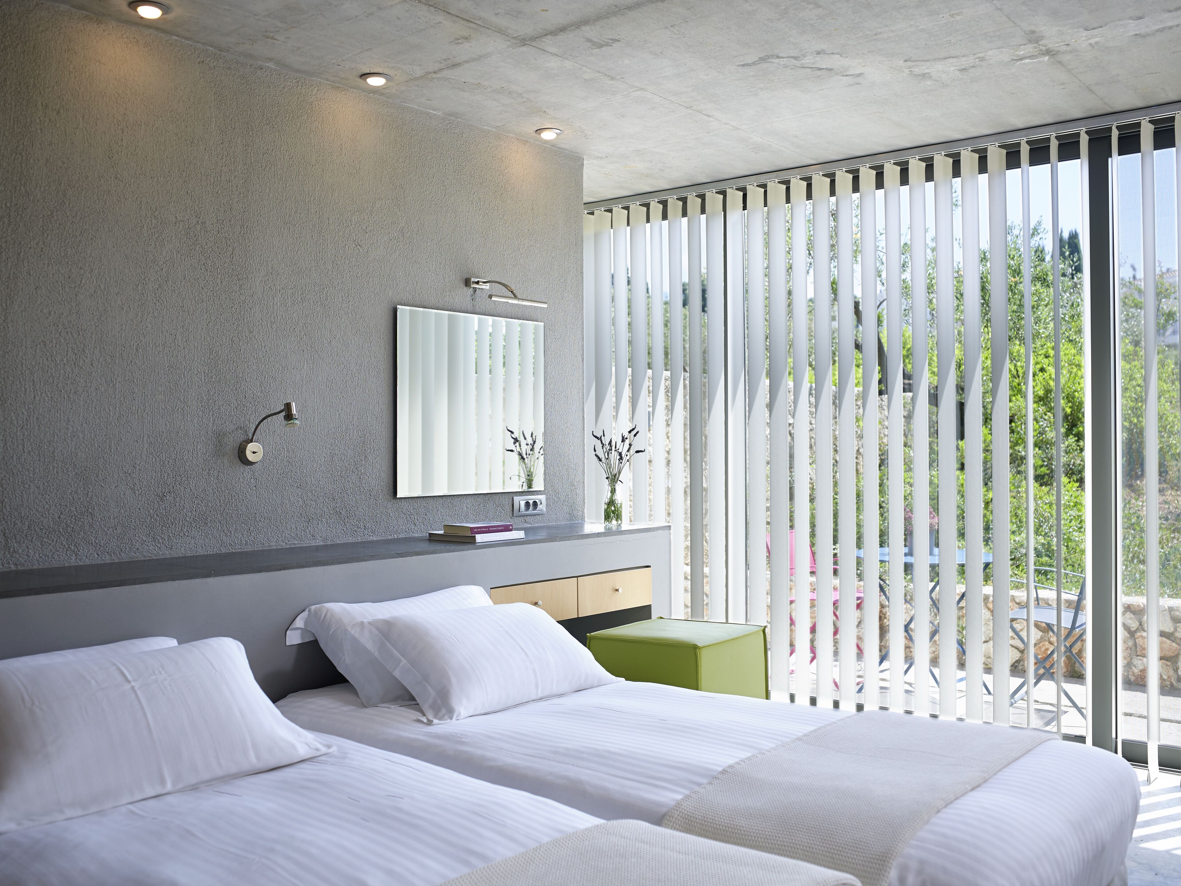 A modern hotel room with two beds, a large window with vertical blinds, a mirror, and a small table with a vase.