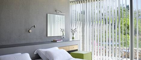 A modern hotel room with two beds, a large window with vertical blinds, a mirror, and a small table with a vase.