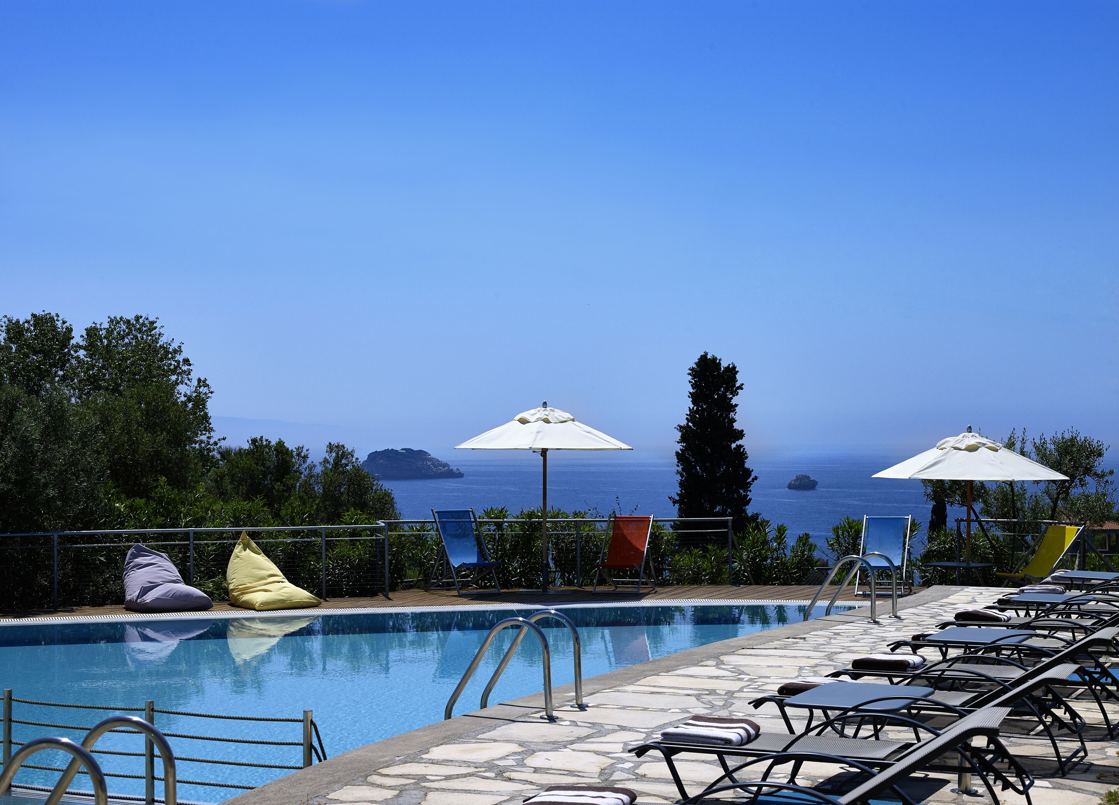 Ein Poolbereich mit Liegestühlen und Sonnenschirmen mit Blick auf ein klares blaues Meer und in der Ferne Inseln.