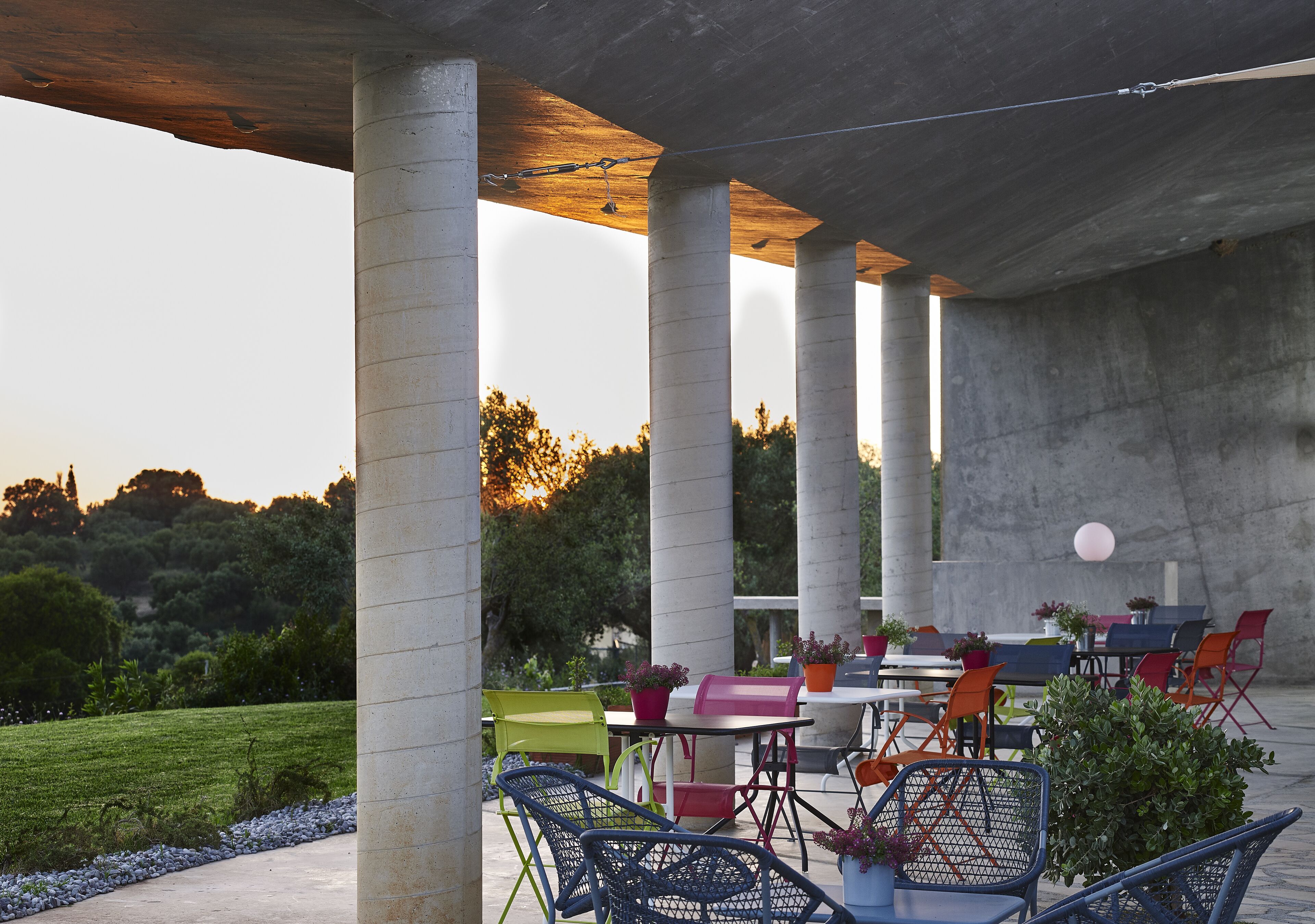 An outdoor patio area with concrete columns, tables, and chairs, surrounded by greenery and a clear sky.