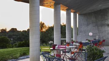 An outdoor patio area with concrete columns, tables, and chairs, surrounded by greenery and a clear sky.