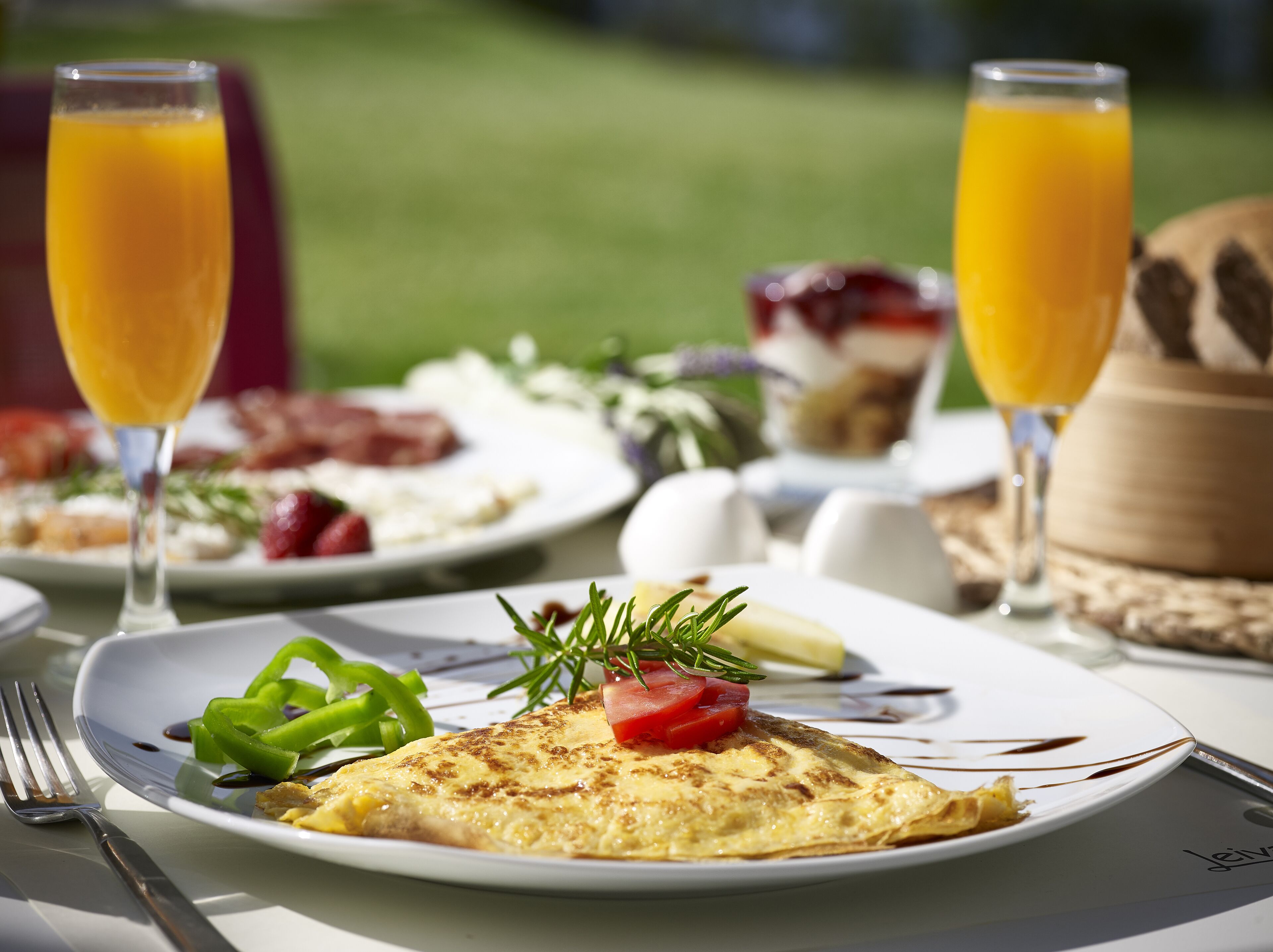 Ein Frühstückstisch mit einer Omelett, grünen Paprika und Tomaten auf einem Teller, dazu zwei Gläser Orangensaft.