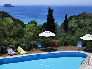 Piscina con vista al mar, rodeada de vegetación y equipada con tumbonas y sombrillas.
