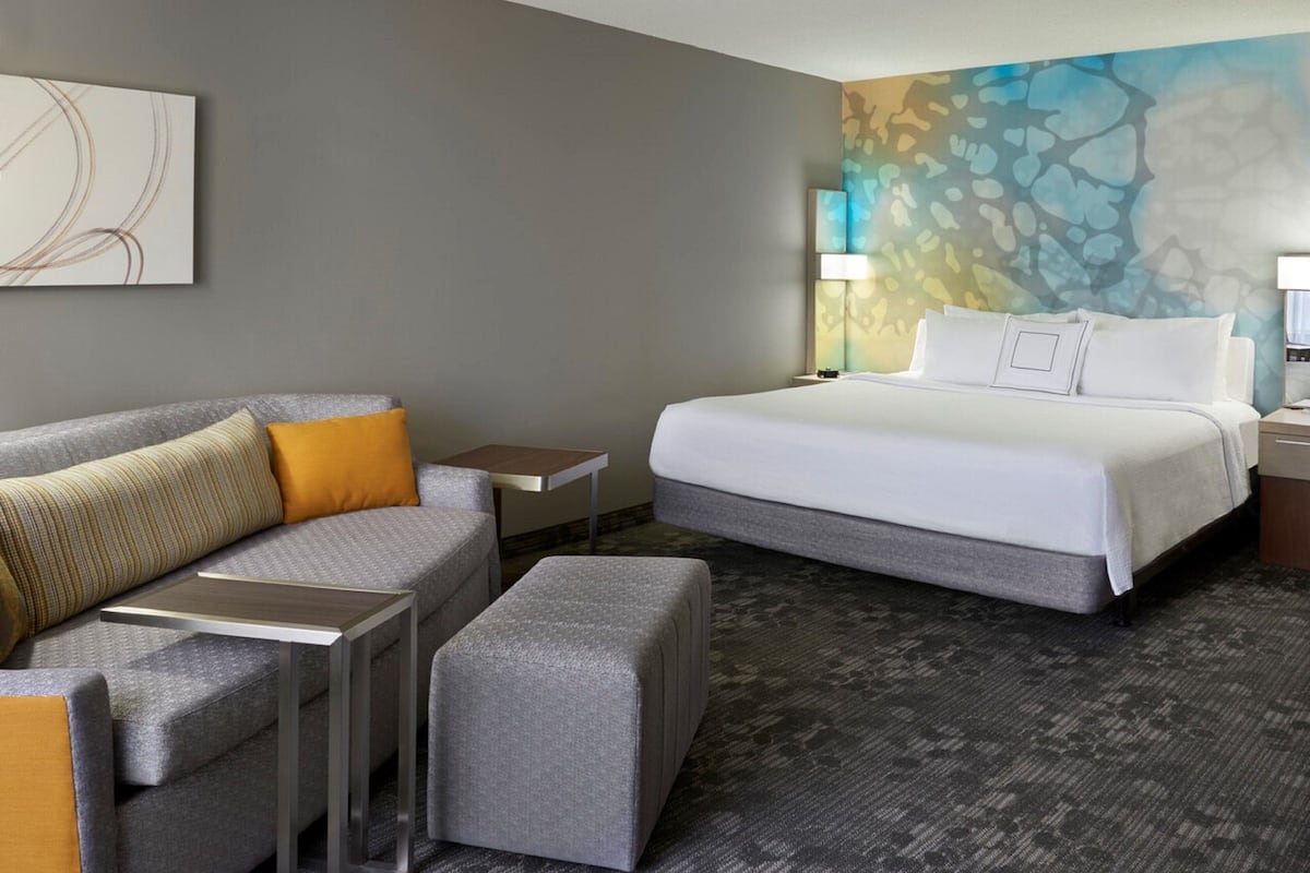 A hotel room with a grey sofa, a matching ottoman, a bed with white linens, and a colorful abstract wall mural.