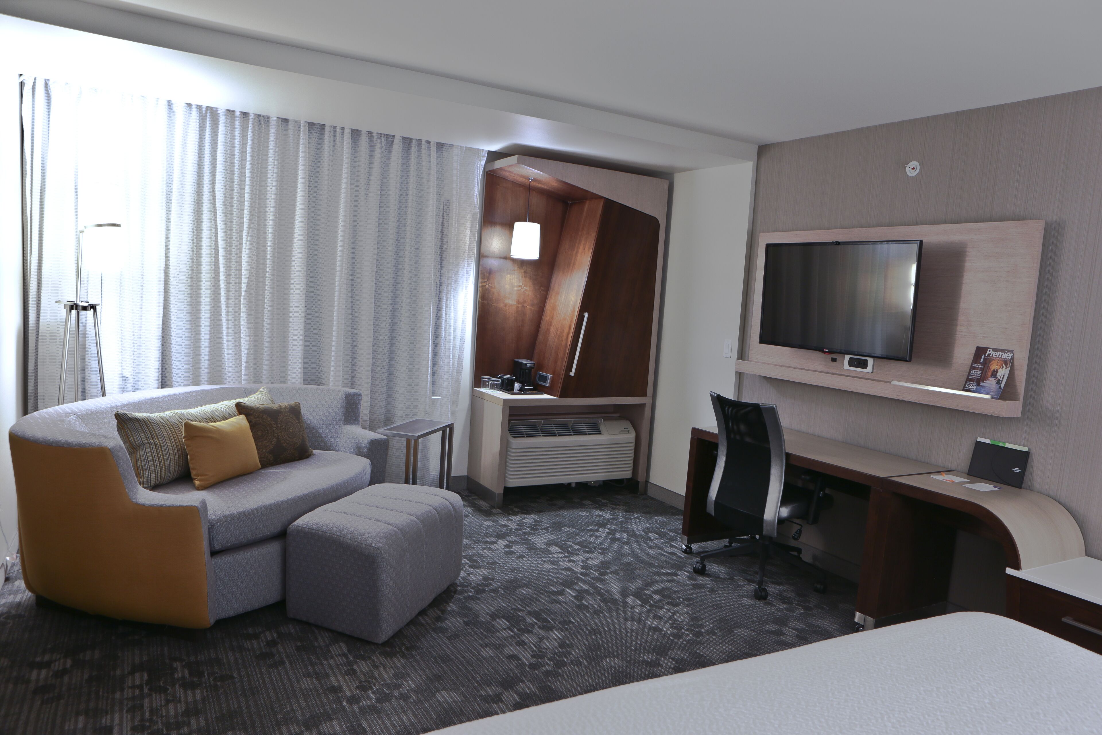 A modern hotel room with a sofa, a desk, a television, and a large window with curtains.