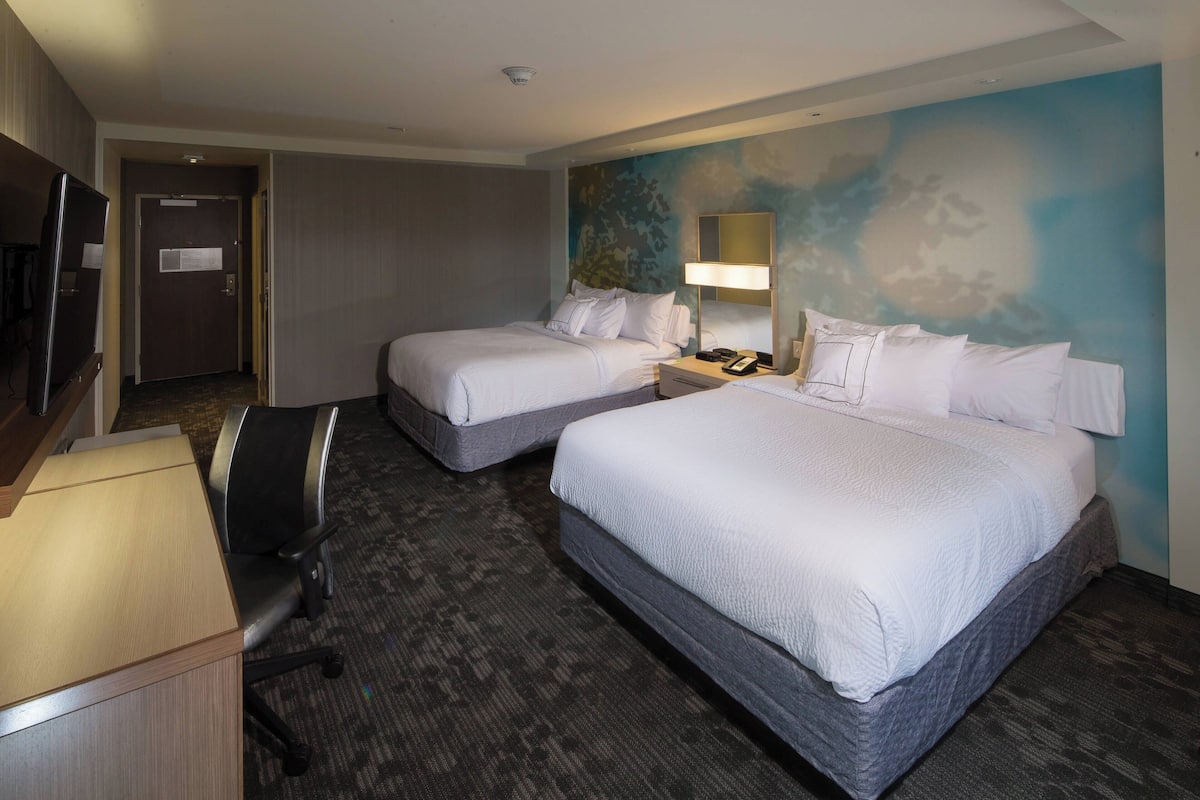 A hotel room with two beds, a desk, a chair, a TV, and a wall with a cloud mural.