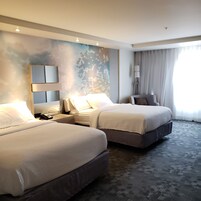 A hotel room with two beds, a large mirror, a bedside table with a phone, a lamp, and a view of the outdoors through sheer curtains.