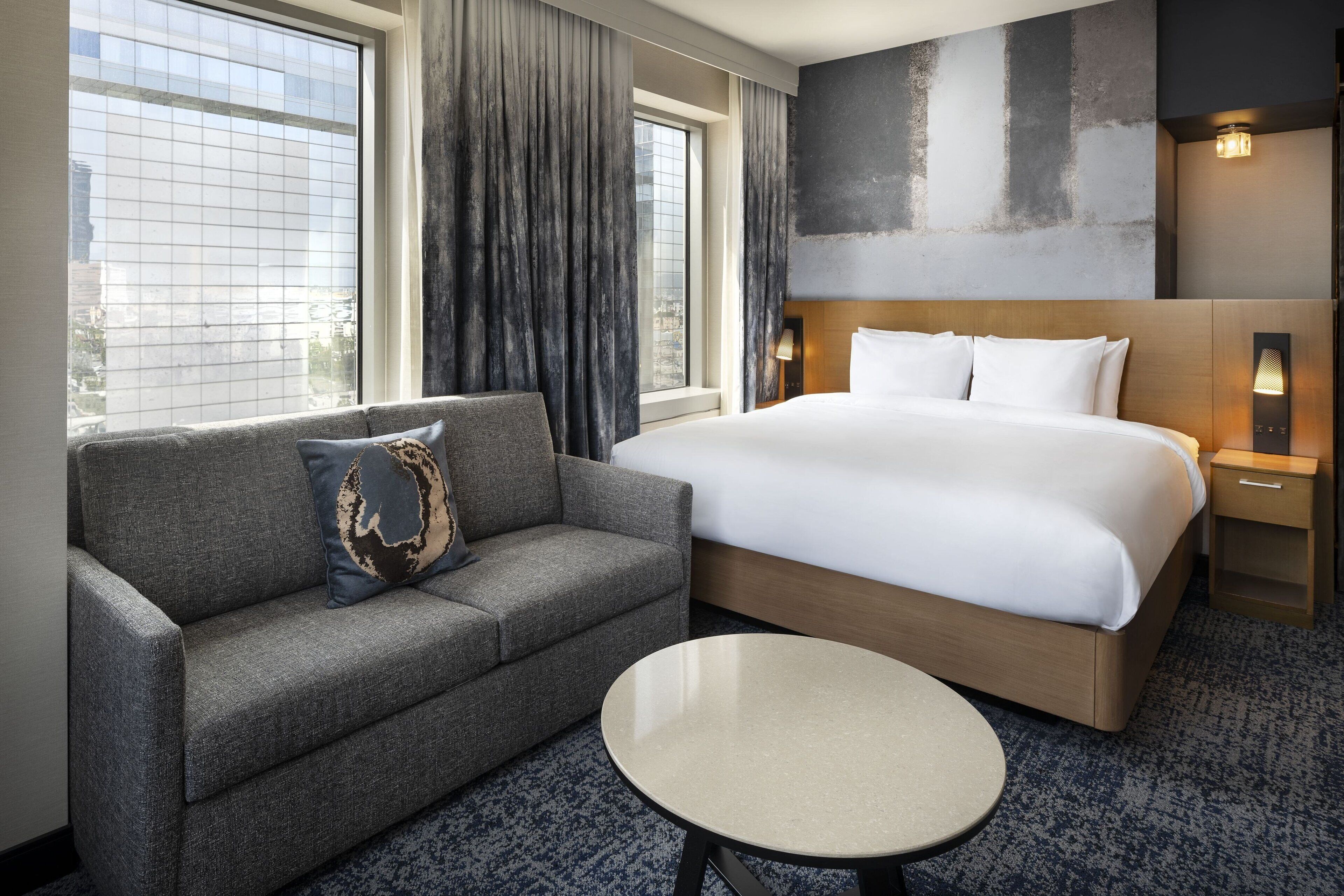 Quarto de hotel com uma cama grande, um sofá, uma mesa redonda e vista para a cidade pela janela.