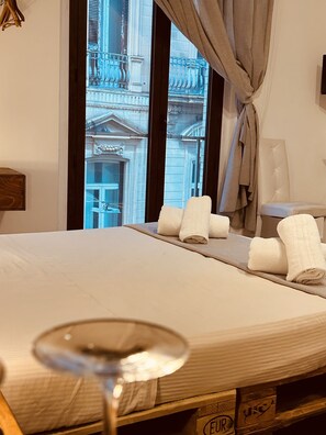 Une chambre d’hôtel avec un lit, une fenêtre avec des rideaux, une vue sur un immeuble et un verre à vin posé sur le sol.