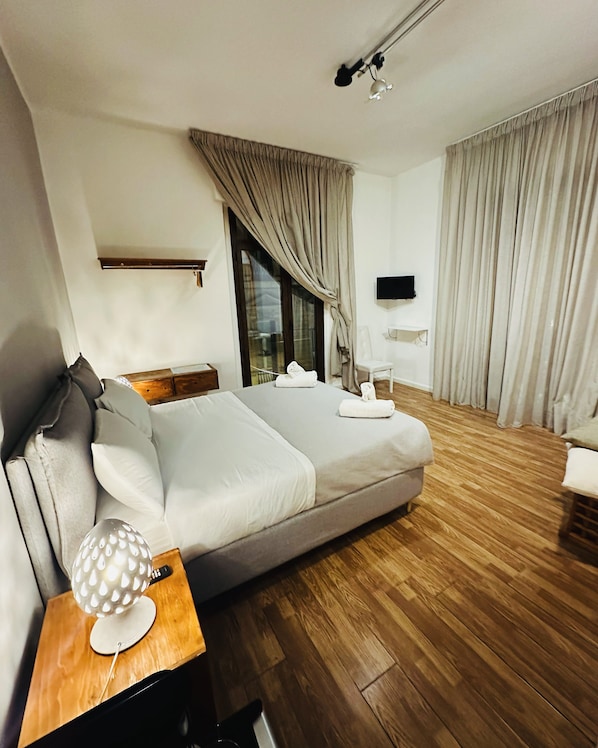 A modern hotel room with a large bed, wooden flooring, a flat-screen TV, and a window with curtains.