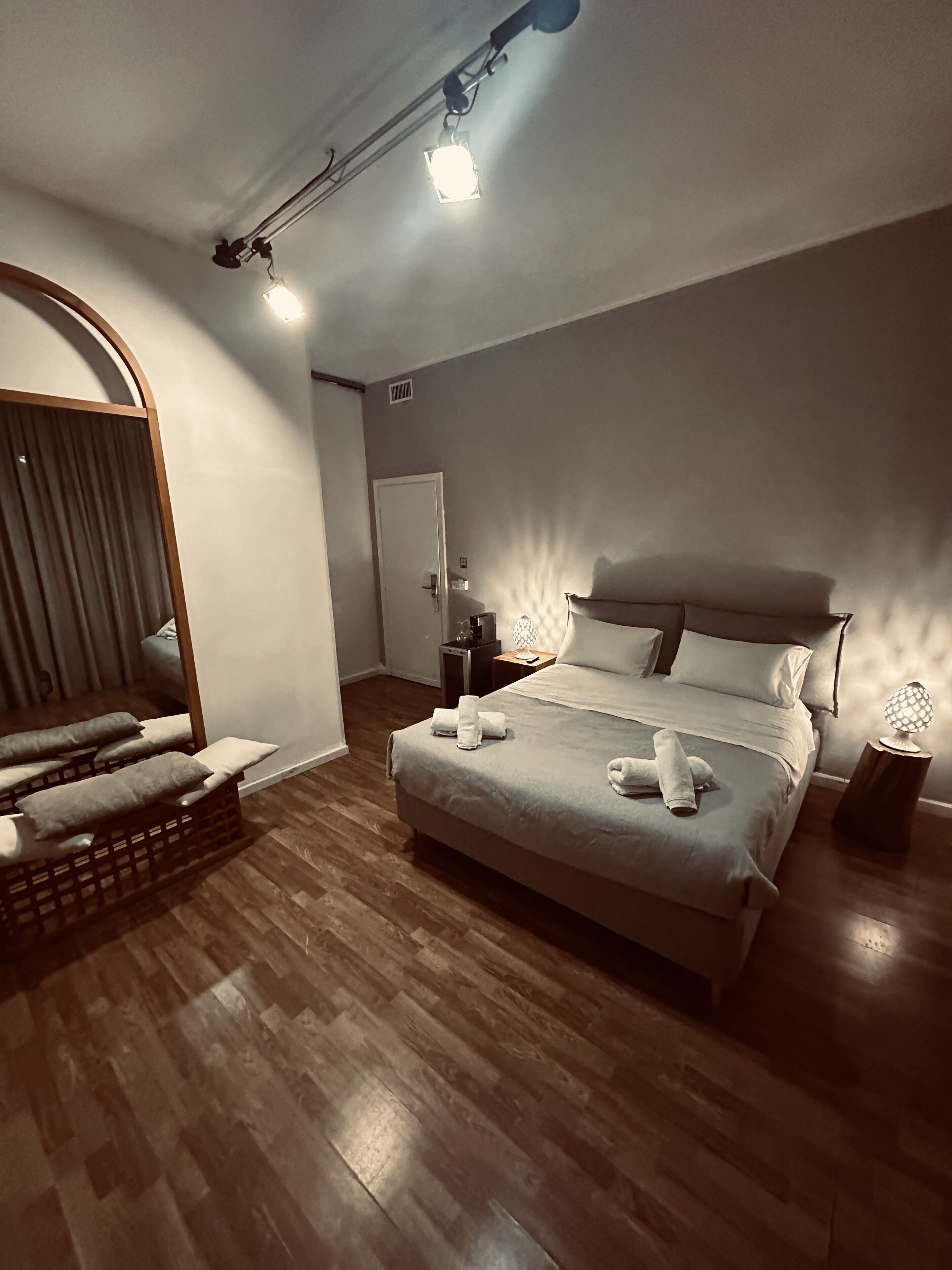 Un dormitorio moderno con una cama grande, suelo de madera, un espejo y una mesita de noche con lámpara.