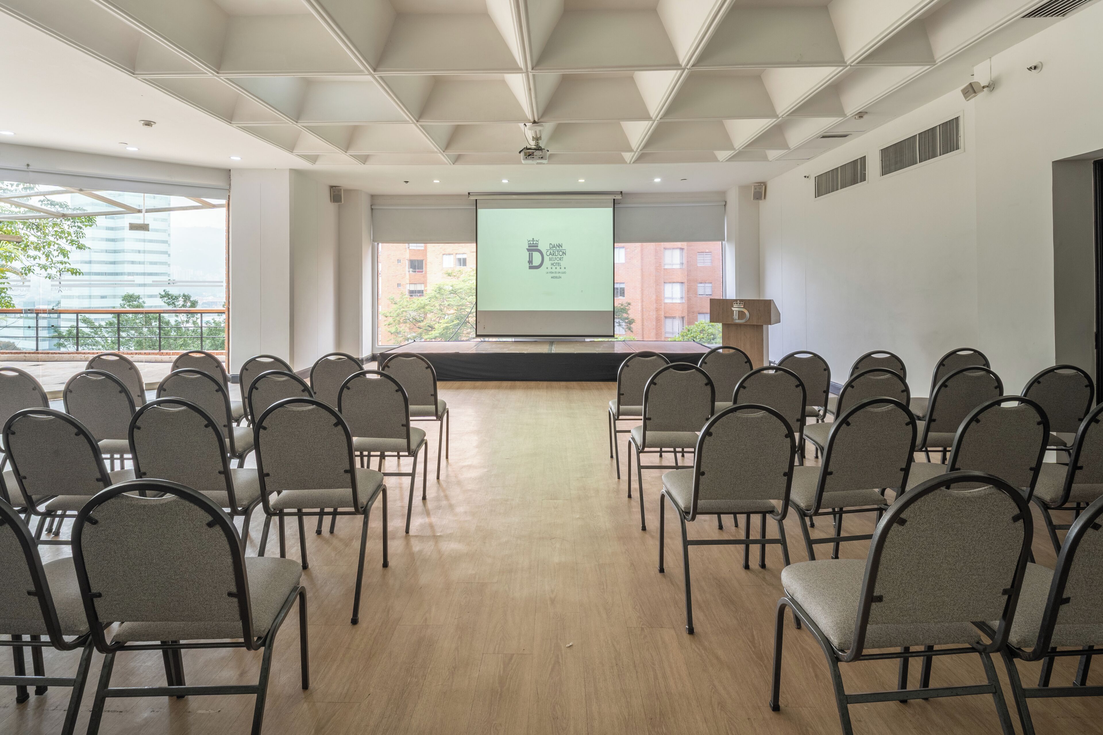 Ein Konferenzraum mit Reihen von Stühlen, die auf einen Bildschirm zeigen, einem Rednerpult und Blick auf Gebäude im Freien.