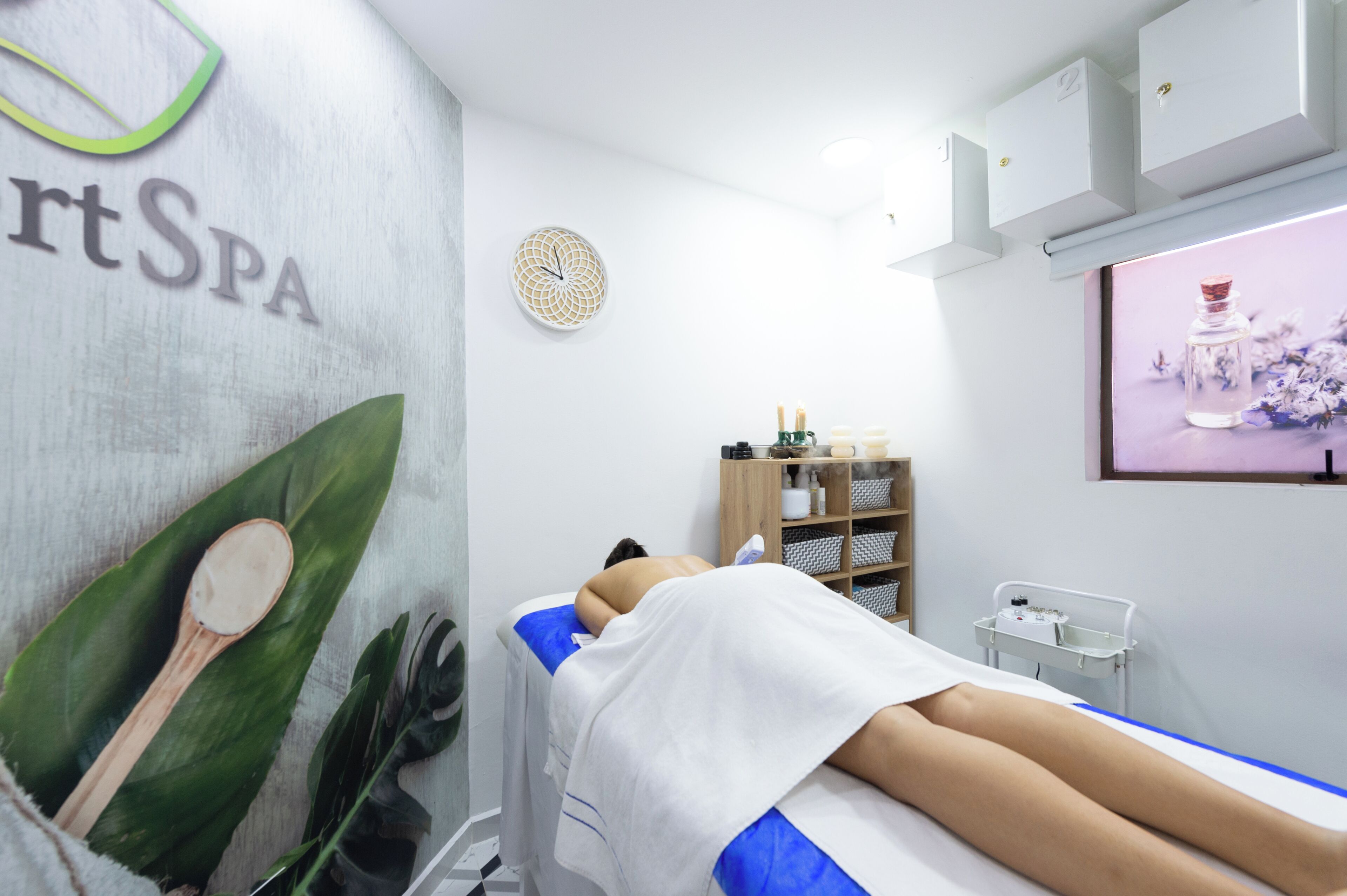 A spa room with a massage table, a wall-mounted shelf with bottles, and a decorative wall piece.