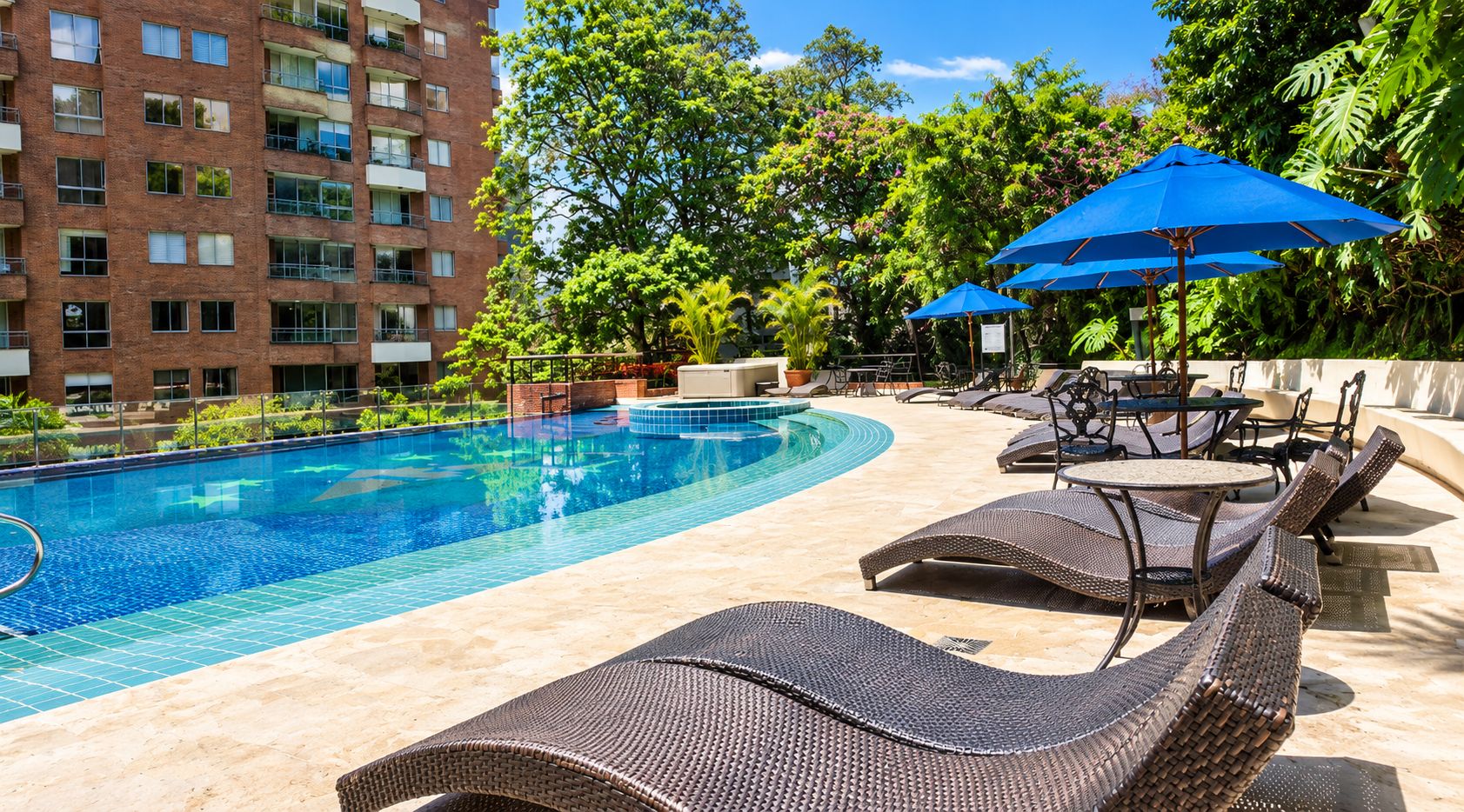 Una piscina con sillas de descanso y sombrillas, rodeada de follaje y un edificio de ladrillo.