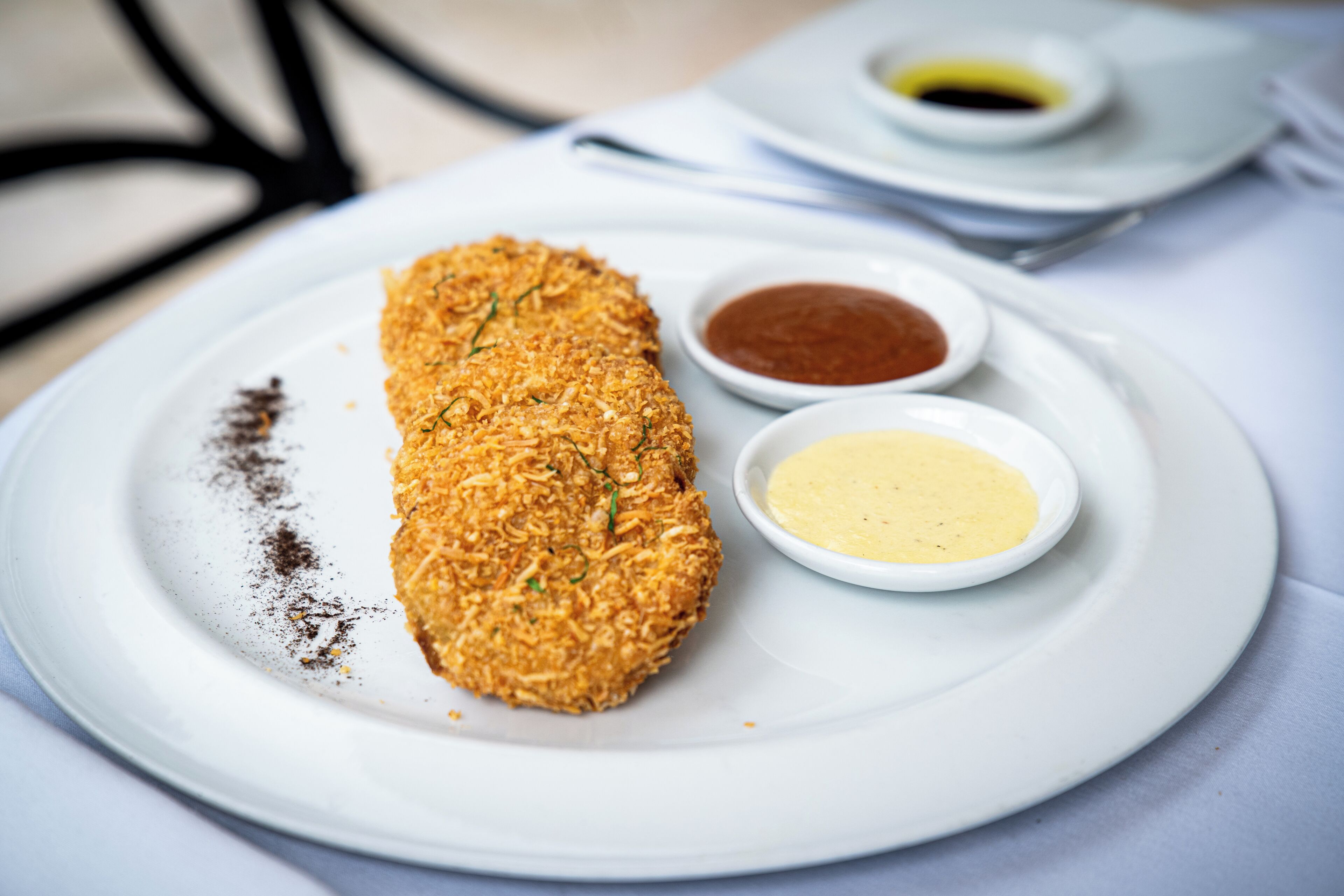 A plate with a breaded item, two dipping sauces, and a small bowl of oil.