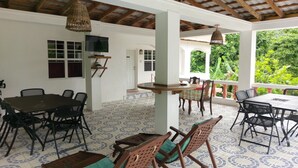 A covered outdoor dining area with multiple tables and chairs, a television mounted on a wall, and a view of lush greenery outside. - Polish Princess Guest House (Port Antonio)