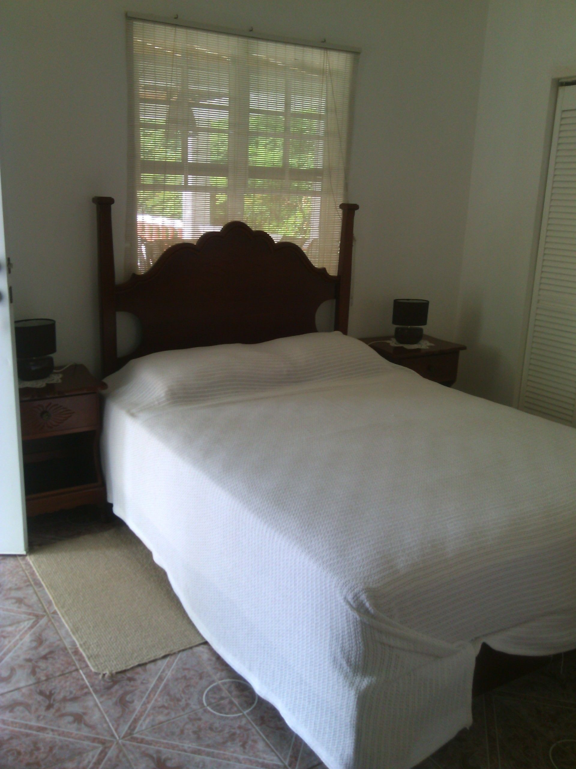 A bedroom with a wooden bed, a nightstand with a lamp, and a window with blinds.