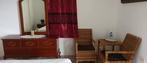A room with a bed, a wooden dresser with drawers, a mirror, a wicker chair, a small table with a water bottle, and a red and black patterned curtain.