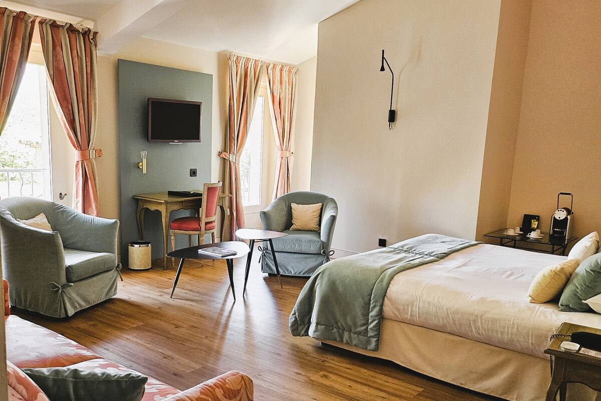 Une chambre d’hôtel comprenant un lit, deux fauteuils, un canapé, une petite table, un téléviseur et une fenêtre avec des rideaux.