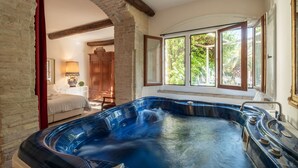 A hot tub in a room with a rustic stone wall, wooden beams, and a window with a view of greenery. - Corte Cristina (Quartucciu)