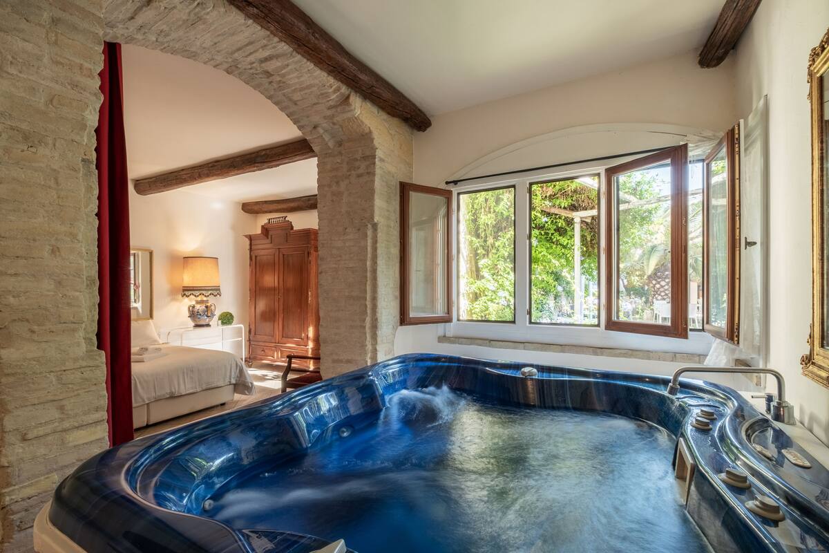 Una bañera de hidromasaje en una habitación con pared de piedra rústica, vigas de madera y ventana con vista a zonas verdes.