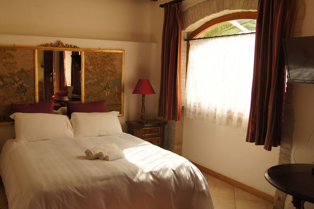 Un dormitorio con una cama grande, un espejo, una mesita de noche, una lámpara y una ventana con cortinas.