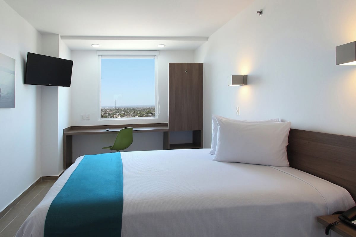 A hotel room with a bed, a desk, a chair, a TV, and a window with a city view.