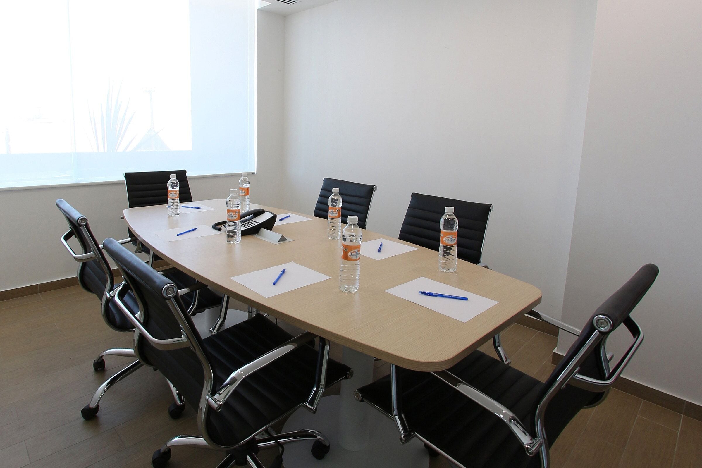 A conference room with a long table, six black chairs, water bottles, pens, and a phone.