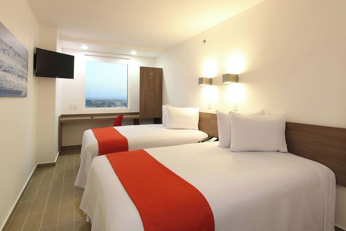 A hotel room with two beds, a TV, a desk, and a window with a city view.