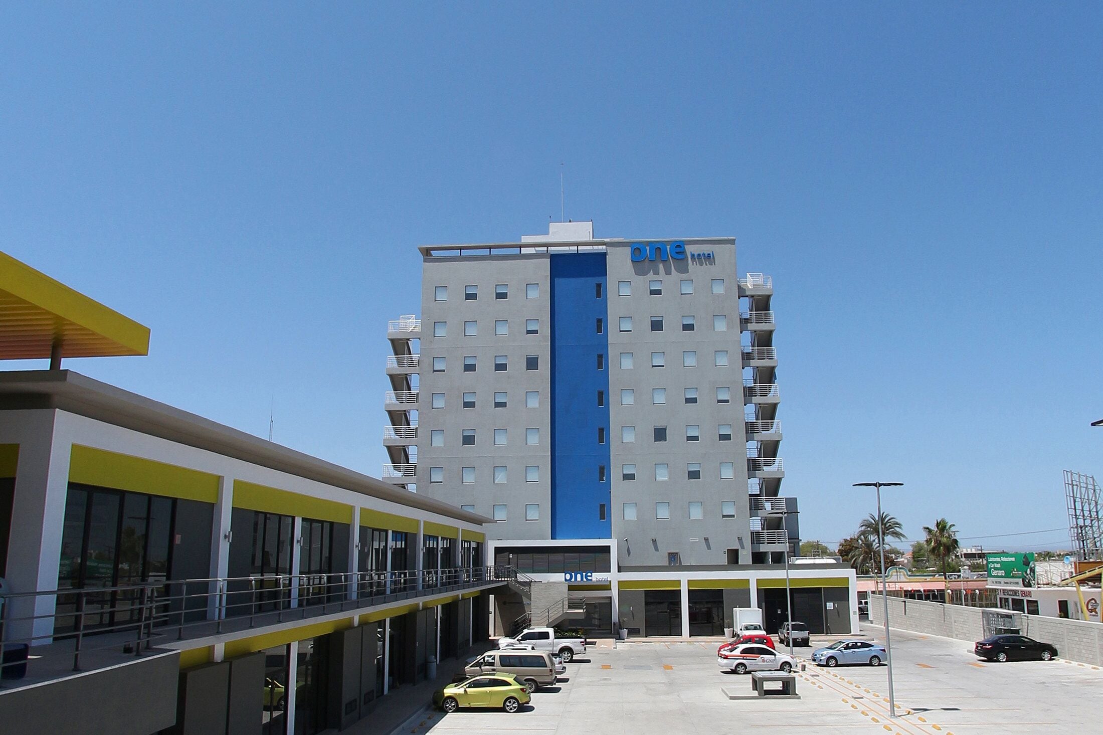 Ein modernes Hotel mit einem auffälligen, blauen, vertikalen Streifen, mehreren Stockwerken und einem Parkplatz mit einigen Autos.