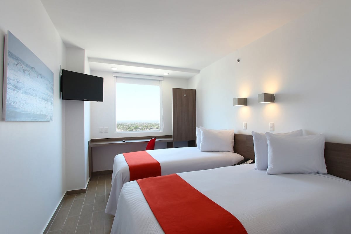 A hotel room with two beds, a TV mounted on the wall, a window with a view, and a desk with a chair.