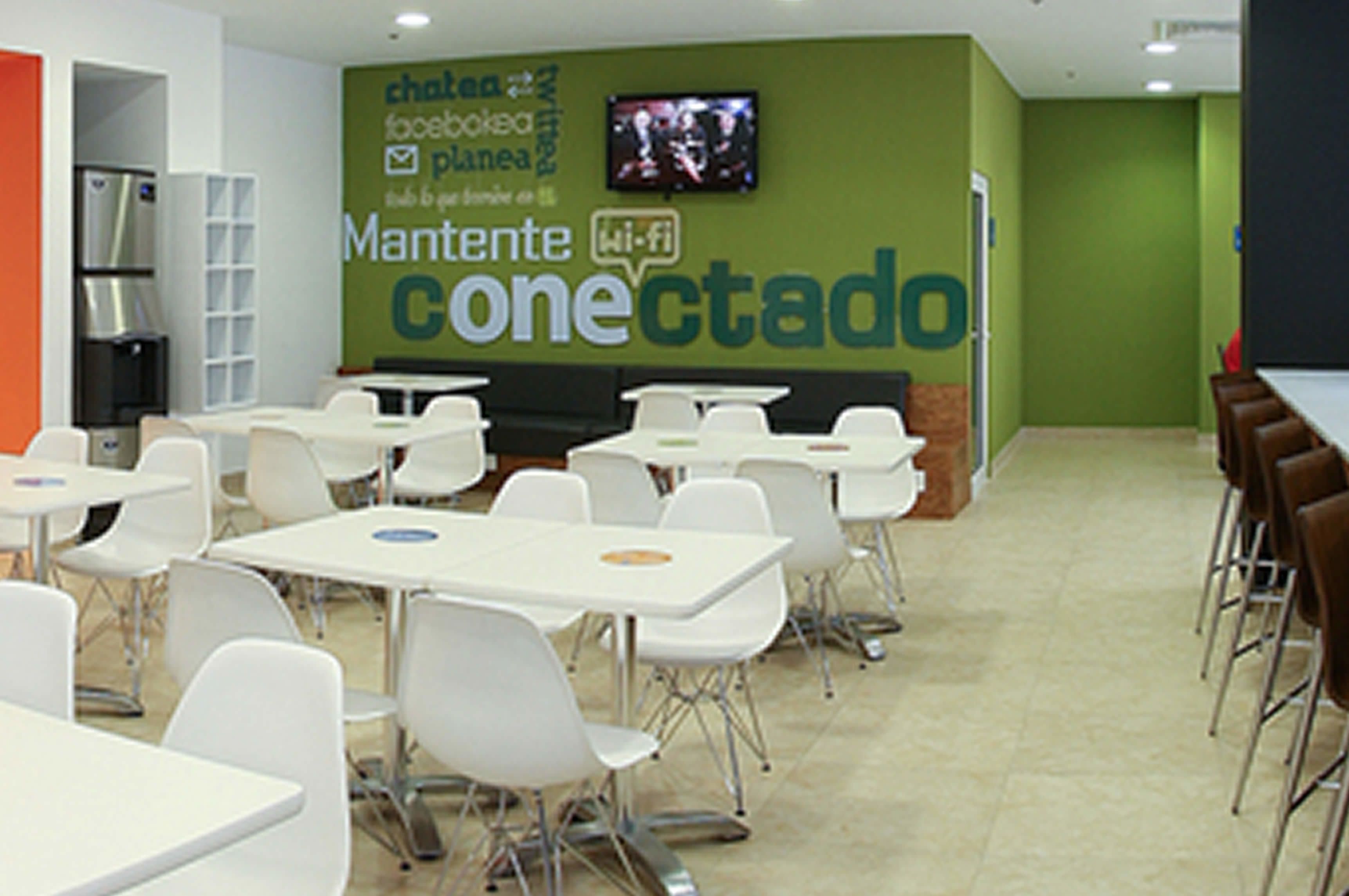 Un comedor con mesas y sillas blancas, una pared verde con el texto 'Mantente conectado' y un letrero de Wi-Fi.