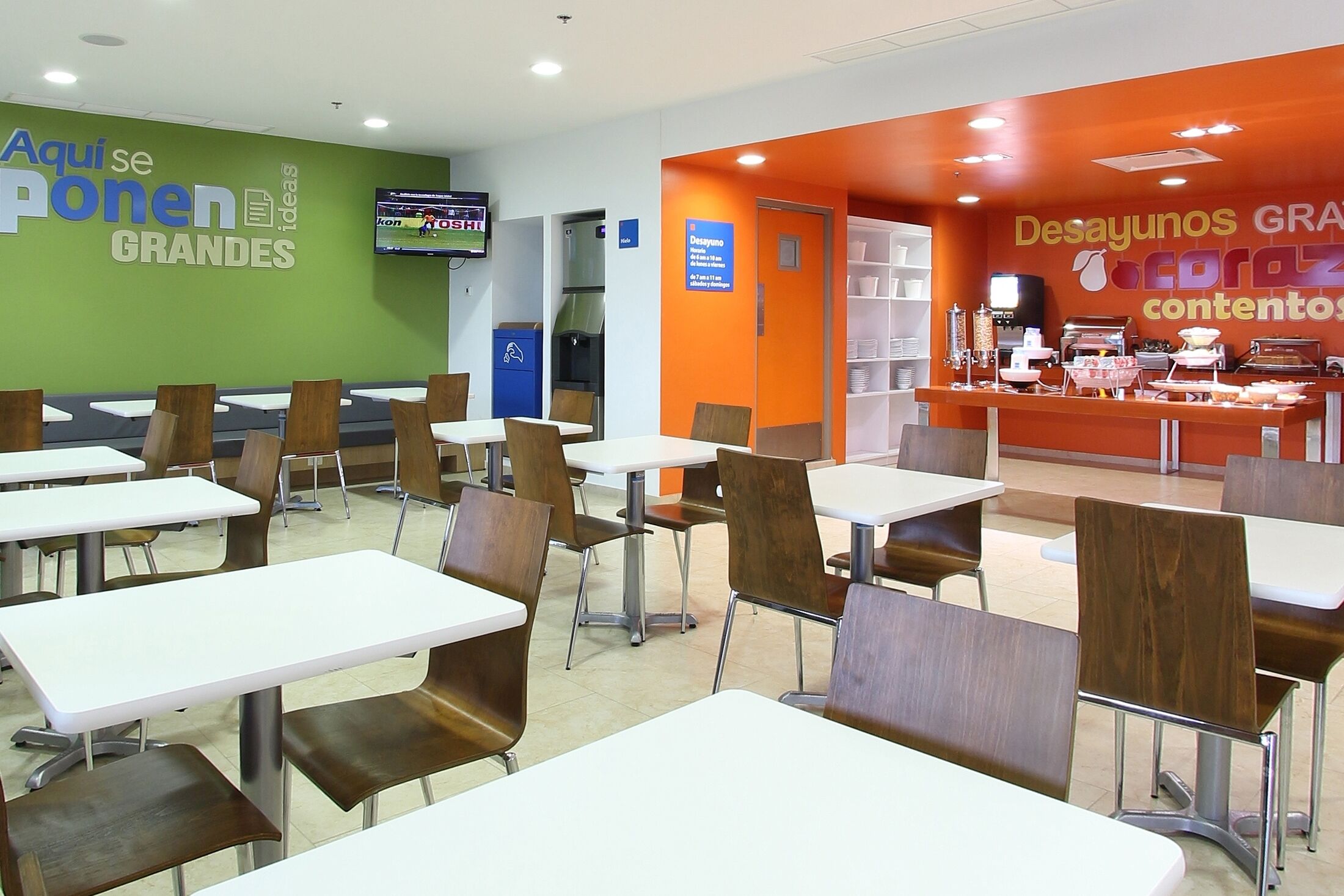 Eine Cafeteria mit weißen Tischen und braunen Stühlen, einem Fernseher an der Wand und einer Theke mit Speisen.