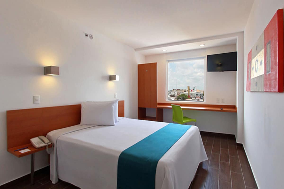 A hotel room with a bed, bedside table, phone, and a window with a city view.