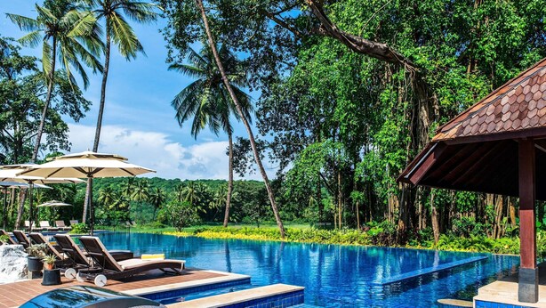 A swimming pool with lounge chairs and umbrellas, surrounded by lush greenery and palm trees. - Novotel Goa Resort & Spa Candolim (Candolim)