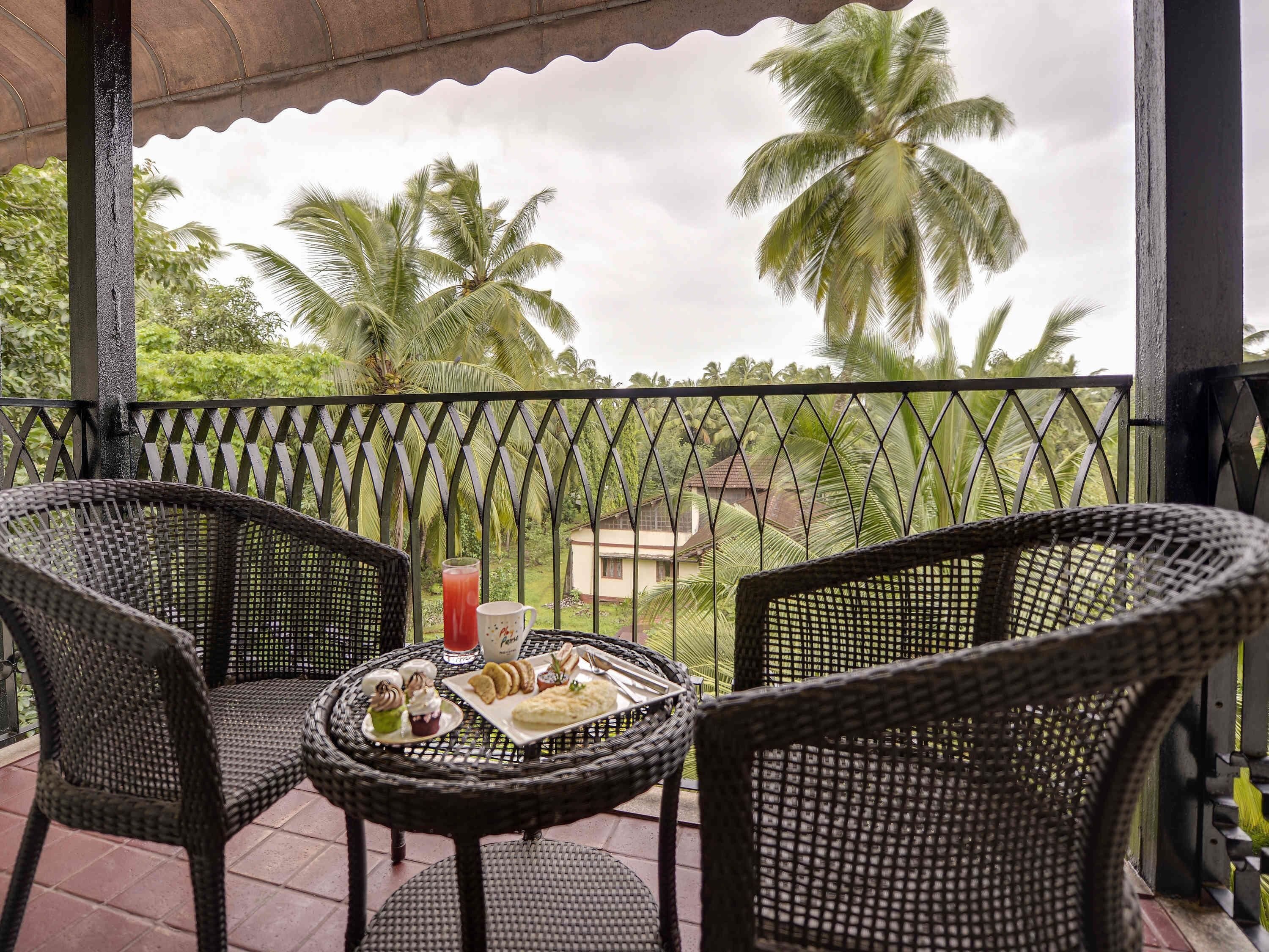 A covered outdoor seating area with wicker chairs and a table set with food and drinks, overlooking palm trees and a distant building.