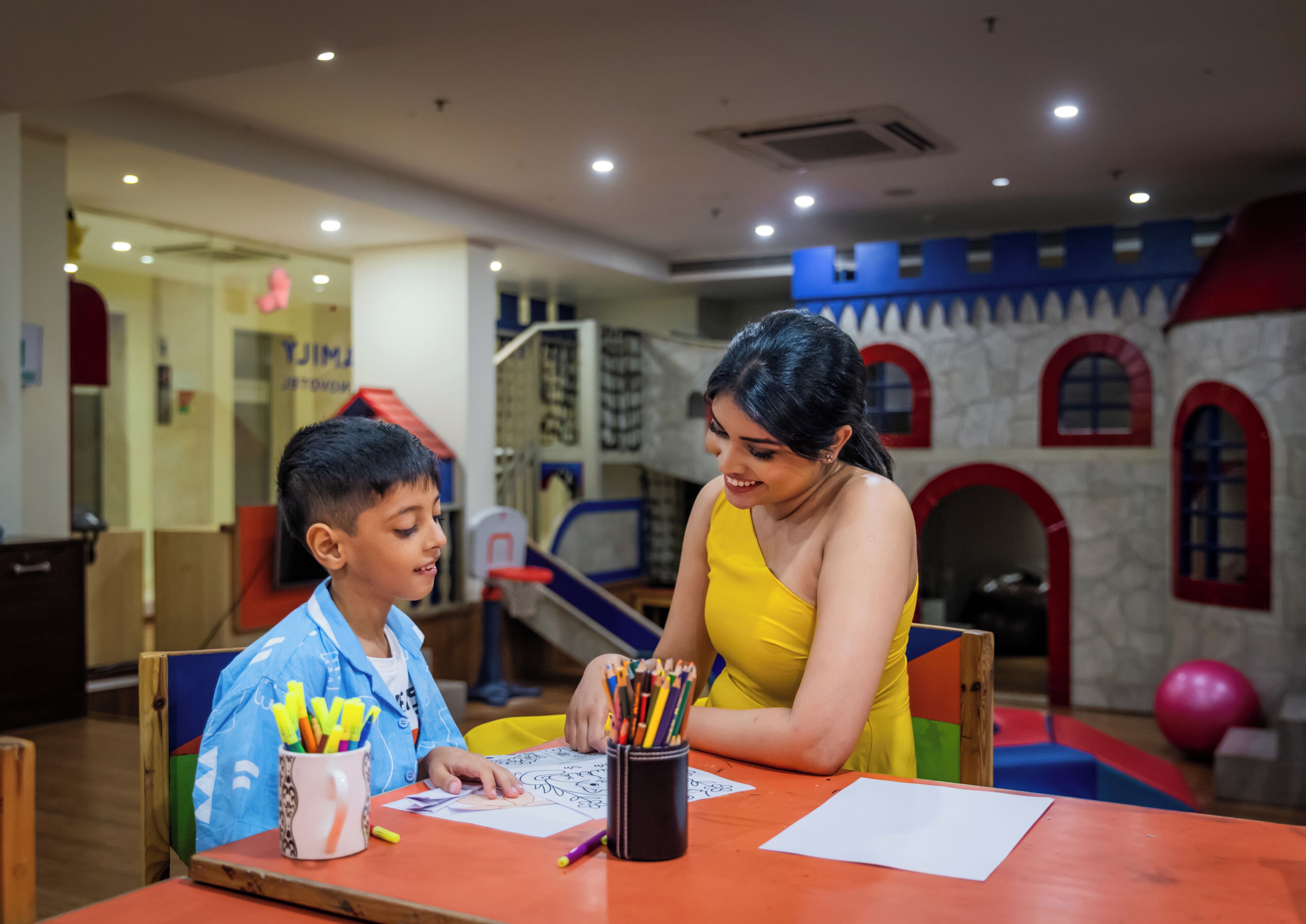 Un enfant et un adulte sont assis à une table, devant du matériel de coloriage, dans une pièce qui comprend un espace de jeux et un mur décoré sur le thème d’un château.