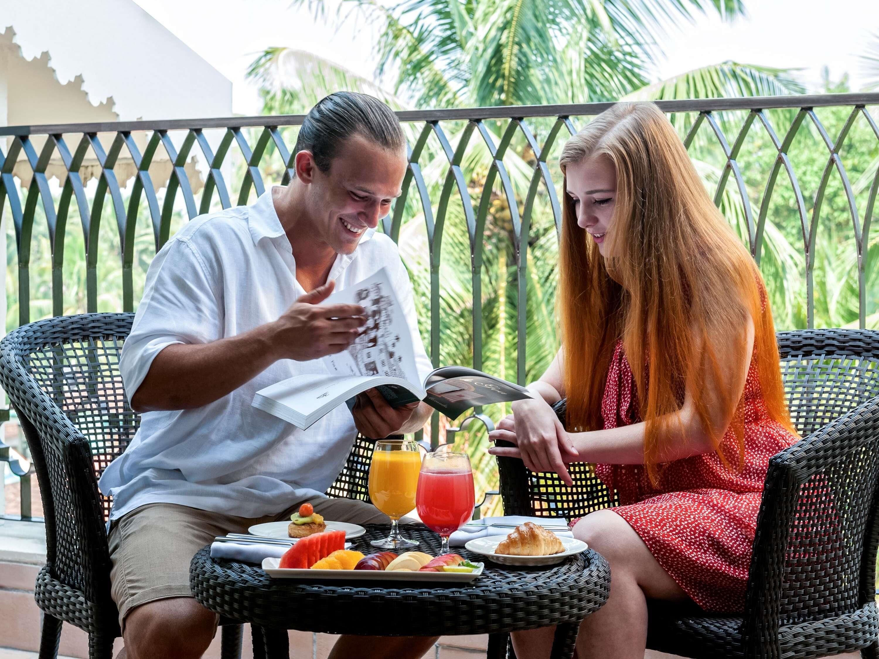 Dos personas sentadas en una mesa con un menú, bebidas y un plato de comida en un balcón con barandilla de metal y follaje al fondo.