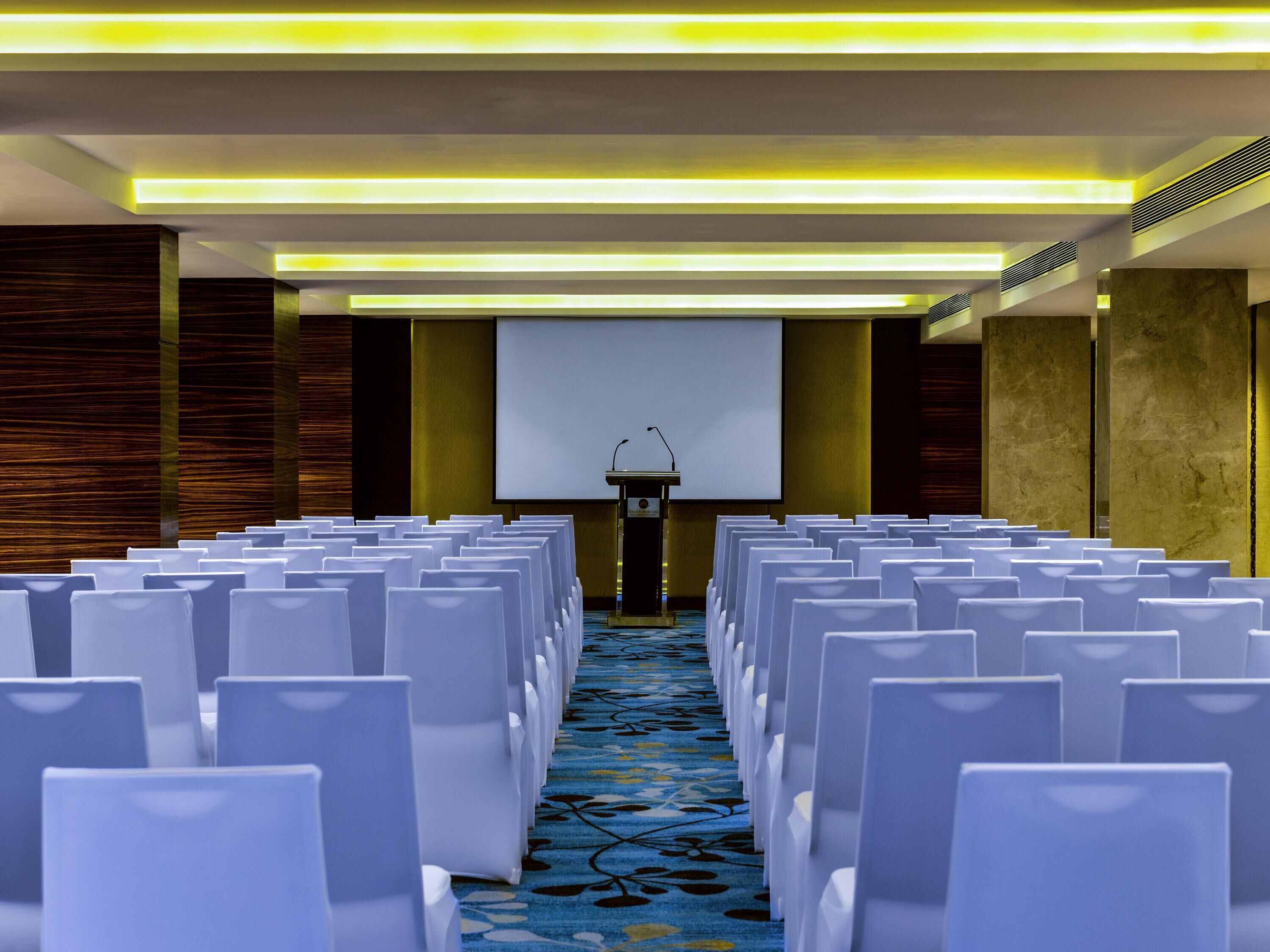 A conference room with rows of white chairs, a podium, and a projector screen.