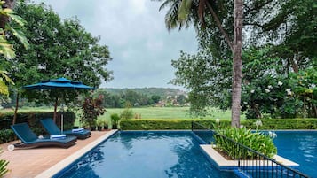 Relax by the serene pool surrounded by lush greenery and palm trees, with lounge chairs and a shaded area for unwinding.