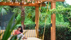 A wooden swing chair with yellow and gray stripes, suspended by ropes, in a lush garden with greenery and a wooden pergola overhead. - Novotel Goa Resort & Spa Candolim (Candolim)