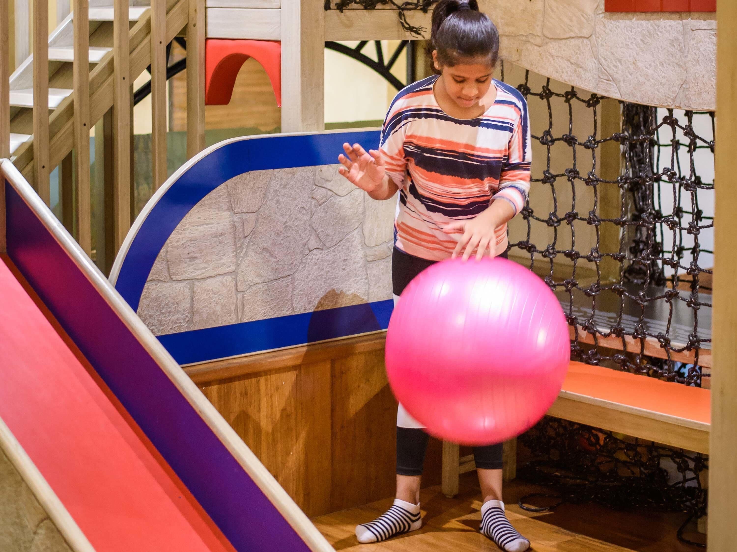 A person playing with a pink ball in a play area with a slide and netted climbing structure.