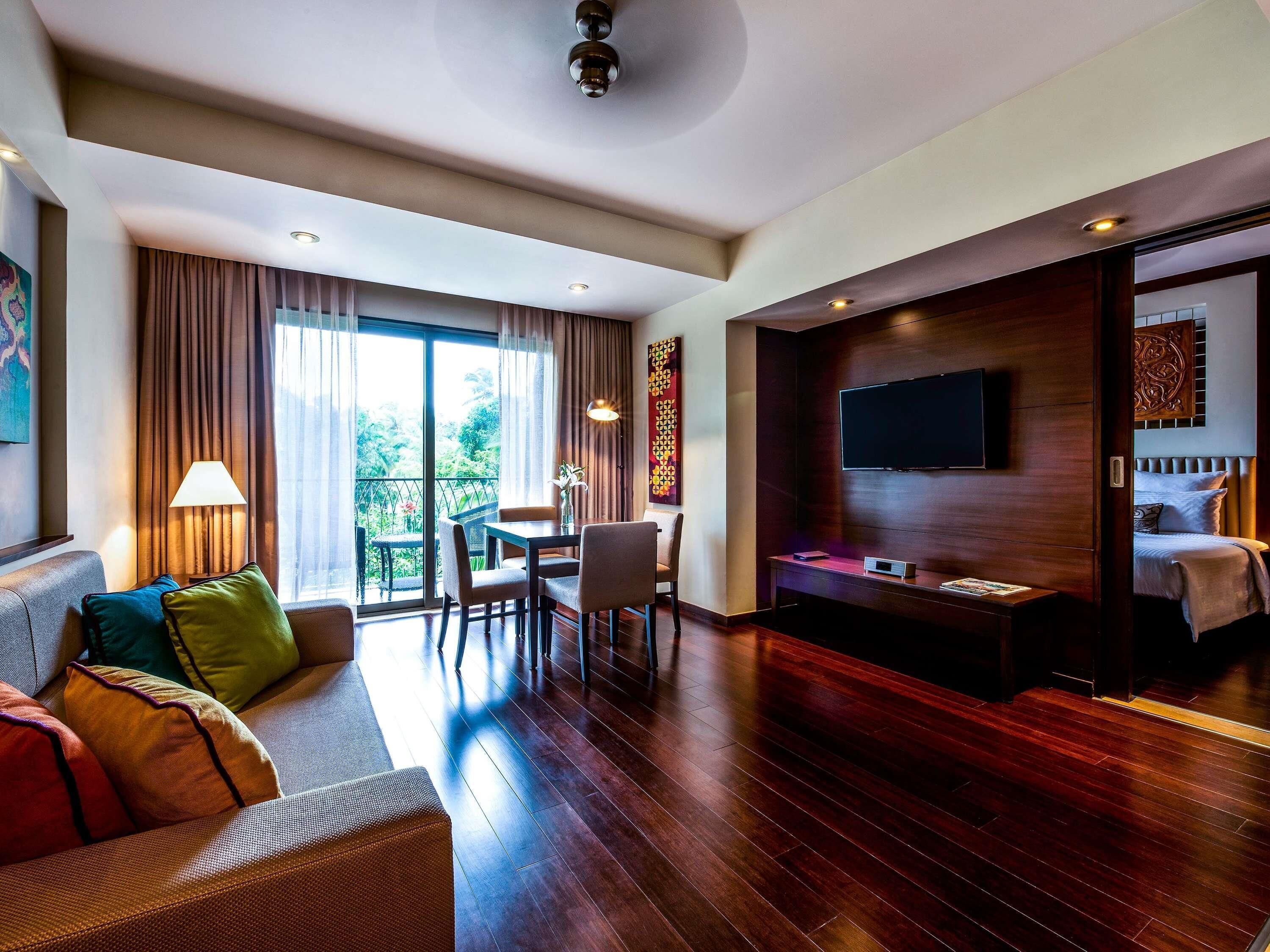 Ein Hotelzimmer mit Sofa, Esstisch, Fernseher und einem Balkon mit Vorhängen.