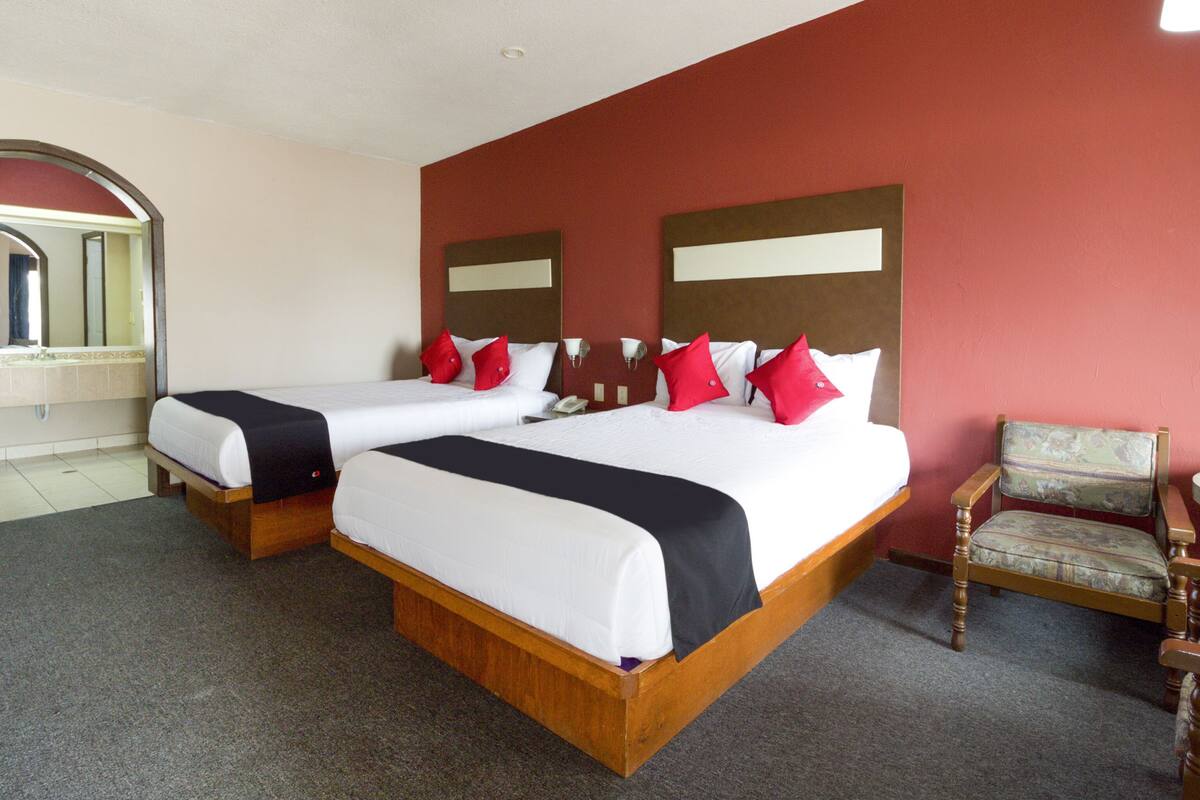 A hotel room with two beds, a wooden headboard, a red accent wall, a wooden chair, and a bathroom with a mirror and sink.